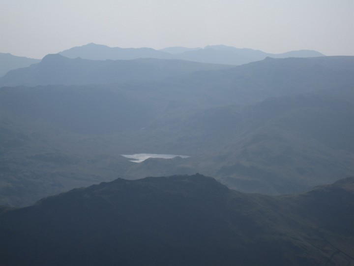 IMG_3478.JPG - View acoss to Helm Crag (foreground) and Easdale Tarn.  High Raise (?) off to the right.  Langdale Pikes (?) and beyond Bow Fell (?) off to the left.  All peaks I'd walked over the last few days.  It was a pity it was hazy and into the sun.